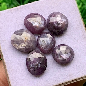 Peut inclure: Un ensemble de six pierres précieuses violettes à facettes de forme ovale avec une finition polie. Chaque pierre présente des inclusions naturelles et des nuances variées de violet profond et de magenta, disposées en groupe sur une surface texturée.