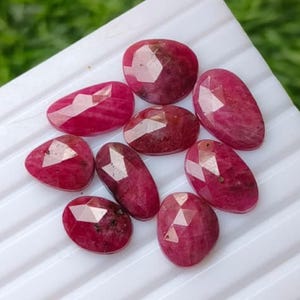 May include: A collection of nine faceted, deep pinkish-red rubies with irregular oval and rounded shapes. Each gemstone features a polished, multi-faceted surface that reflects light, showcasing a vibrant, saturated colour and natural internal inclusions.