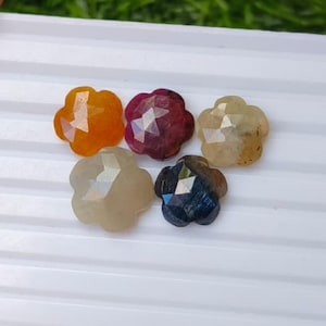 Puede incluir: Cinco piedras preciosas facetadas talladas en forma de flor, dispuestas en grupo. Las piedras son translúcidas y muestran colores distintos, incluyendo naranja, rojo oscuro, amarillo pálido, gris claro y azul oscuro con inclusiones.