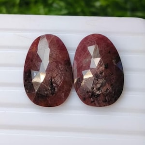 Könnte beinhalten: Zwei ovale, facettierte Edelsteine in einem tiefen Rotbraun. Jeder Stein hat eine polierte Oberfläche mit sichtbaren inneren Einschlüssen und dunklen Mineralstreifen, die das Licht auf den geometrischen Facetten der Oberseite reflektieren.