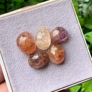 Puede incluir: Cinco piedras preciosas facetadas de forma ovalada dispuestas en un grupo sobre una almohadilla de exhibición de color gris claro texturizado. Las piedras muestran una variedad de colores translúcidos, incluyendo tonos naranja, melocotón, marrón claro y violeta.
