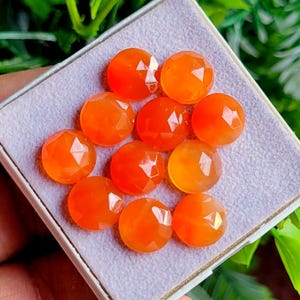 May include: A collection of ten faceted, round, orange gemstones displayed in a white box. The gemstones have a polished, reflective surface, and are arranged closely together. The background is a soft, blurred green.