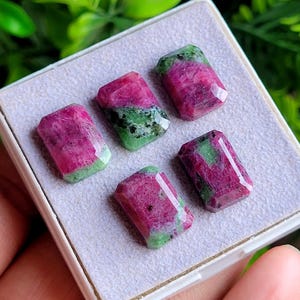 May include: Five rectangular faceted gemstones featuring a mix of vibrant magenta-pink and deep forest green mineral inclusions. The stones are arranged in a display box with a light gray textured foam insert, showcasing their polished surfaces.
