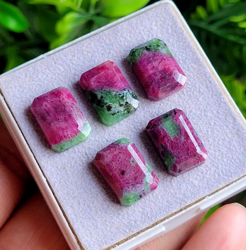 May include: Five rectangular faceted gemstones featuring a mix of vibrant magenta-pink and deep forest green mineral inclusions. The stones are arranged in a display box with a light grey textured foam insert, showcasing their polished surfaces.