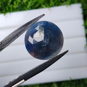 Può includere: Una pietra preziosa blu rotonda e sfaccettata tenuta da una pinzetta di metallo. La pietra mostra una sfumatura dal blu navy intenso al blu più chiaro con inclusioni interne visibili e una superficie lucida che riflette la luce.