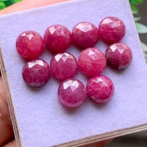 Puede incluir: Un conjunto de diez piedras preciosas redondas, facetadas y de color rojo rosado intenso, dispuestas sobre una almohadilla de exhibición de color violeta claro. Cada piedra presenta un corte geométrico simétrico con múltiples facetas.