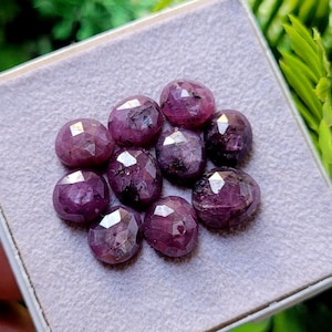 May include: A collection of faceted, deep red-purple gemstones. The stones are oval-shaped and have a polished, reflective surface. They are arranged in a gray, rectangular box, with a few green leaves in the background.
