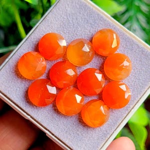 May include: A collection of ten faceted, round, orange gemstones. The gemstones are displayed in a small, square, white-edged tray with a gray interior. The gemstones have a polished, reflective surface.