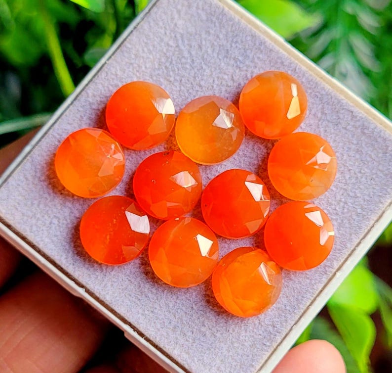 May include: A collection of ten faceted, round, orange gemstones. The gemstones are displayed in a small, square, white-edged tray with a gray interior. The gemstones have a polished, reflective surface.