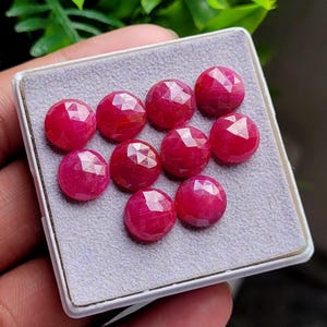 Puede incluir: Una colección de diez piedras preciosas redondas, facetadas y de un vibrante color rojo rosado, dispuestas en una caja de presentación cuadrada con un forro de terciopelo gris claro. Cada piedra presenta un corte geométrico.