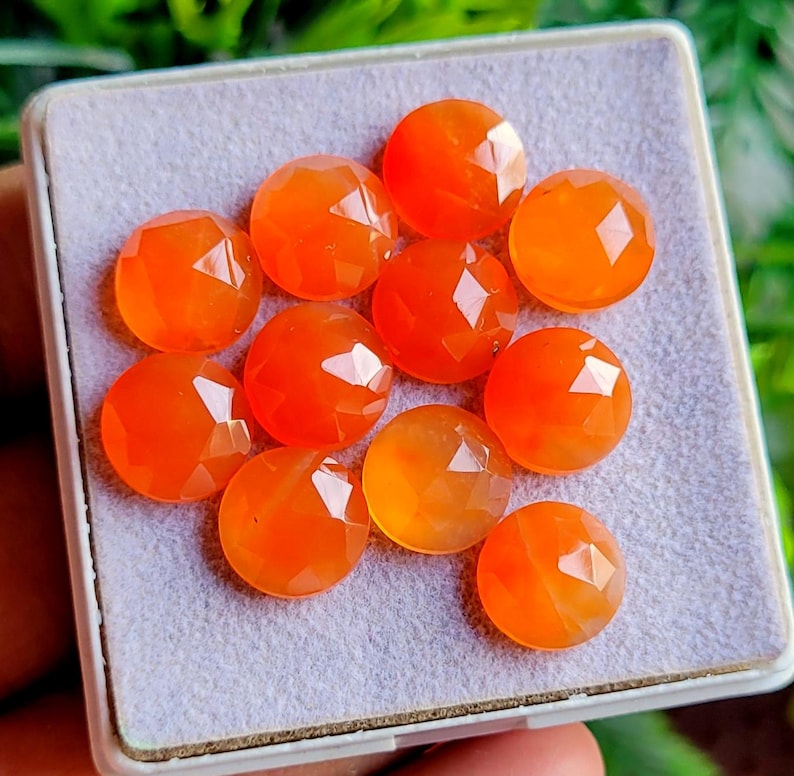 May include: A collection of ten faceted, round, orange gemstones displayed in a white tray with a gray interior. The gemstones have a polished, reflective surface, and are arranged closely together.