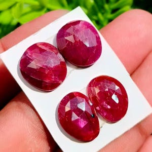 May include: Four faceted, oval-shaped rubies with a deep red colour and a polished finish. The gemstones are arranged on a small white rectangular card, showcasing their reflective surfaces and geometric cut patterns across the top faces.