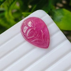 May include: A teardrop-shaped gemstone in a vibrant magenta-pink hue, featuring an intricate, hand-carved floral design on its surface. The stone has a smooth, polished finish and a translucent quality that catches the light, highlighting the delicate petals and stem etched into the centre of the piece.