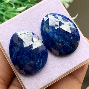 Peut inclure: Deux pierres précieuses facettées bleu profond de forme ovale avec une finition polie. Les pierres présentent une couleur riche et saturée avec des textures internes visibles et des facettes géométriques qui reflètent la lumière.