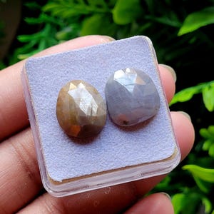 Peut inclure: Deux pierres précieuses à facettes reposant sur un coussinet carré violet clair à l'intérieur d'un boîtier en plastique transparent. L'une est d'une couleur brun doré translucide, tandis que l'autre est gris-bleu semi-transparent et polie.