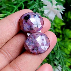 May include: Two faceted, round gemstones with a deep reddish-purple hue. The gems have a polished, reflective surface with multiple facets, and are held in a hand against a backdrop of green foliage and white flowers.