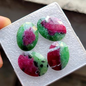 Puede incluir: Cuatro piedras preciosas ovaladas con corte facetado. Las gemas presentan una mezcla de colores, incluyendo rojo intenso, verde vibrante y motas negras. Las piedras se exhiben en una bandeja de plástico blanco.