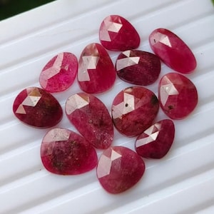 Puede incluir: Una colección de doce rubíes sueltos y facetados en diversas formas ovaladas y redondeadas. Cada piedra preciosa muestra un tono rojo intenso y vibrante con una superficie pulida de múltiples facetas que refleja la luz, dejando ver inclusiones naturales.