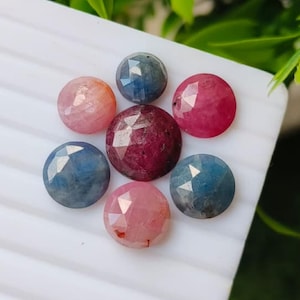 May include: A collection of seven round, faceted gemstones arranged in a cluster. The stones feature a variety of colors including deep red, light pink, and shades of blue, each displaying a geometric cut surface that reflects light across the facets.