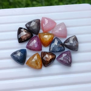 May include: A collection of twelve faceted, triangle-shaped gemstones in various colours including pink, yellow, blue, brown, and dark grey. Each stone features a geometric cut with multiple flat surfaces that reflect light, arranged on a white surface.