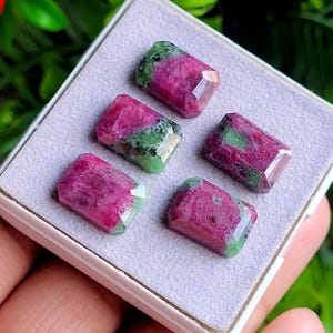 May include: Five rectangular faceted gemstones arranged in a white display box. Each stone features a distinct combination of deep magenta-pink and vibrant green mineral inclusions, showcasing a polished surface with multiple geometric facets.