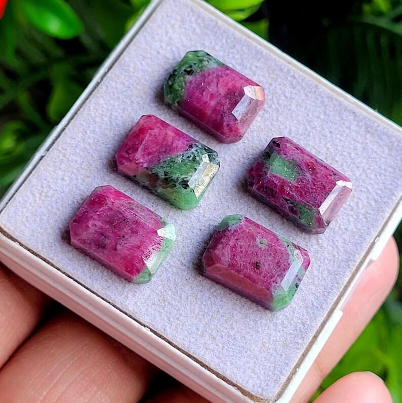 May include: Five rectangular faceted gemstones arranged in a white display box. Each stone features a distinct combination of deep magenta-pink and vibrant green mineral inclusions, showcasing a polished surface with multiple geometric facets.