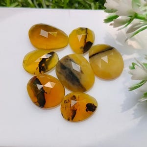 May include: Seven faceted, oval-shaped yellow gemstones with distinct dark brown to black dendritic inclusions. Each stone features a polished, reflective surface with geometric facets that catch the light, displaying a warm golden hue.
