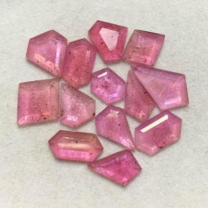 Può includere: Una collezione di dodici pietre preziose rosa sfaccettate con varie forme geometriche, inclusi tagli pentagonali, esagonali e trapezoidali. Ogni pietra mostra una qualità traslucida con inclusioni interne e una superficie lucida e riflettente.
