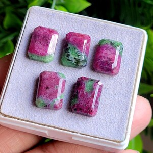 May include: Five rectangular faceted gemstones featuring a mix of vibrant magenta and deep green mineral inclusions. The stones are arranged in a white display box with a soft, light-coloured fabric lining, showcasing their polished surfaces.