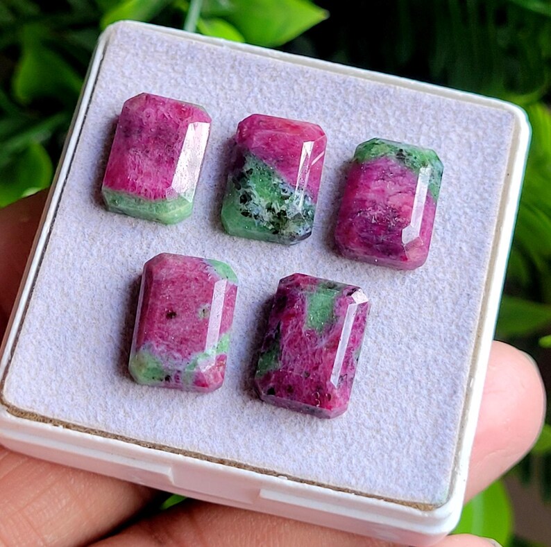 May include: Five rectangular faceted gemstones featuring a mix of vibrant magenta and deep green mineral inclusions. The stones are arranged in a white display box with a soft, light-coloured fabric lining, showcasing their polished surfaces.