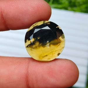 May include: A round, faceted gemstone featuring a gradient of translucent golden-yellow at the bottom transitioning into a deep, opaque black at the top. The stone has a polished, reflective surface with visible internal inclusions.