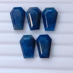 Puede incluir: Cinco piedras preciosas facetadas cortadas en forma de ataúd. Cada piedra presenta un color azul profundo con una superficie pulida y reflectante, además de facetas geométricas distintivas que captan la luz, dispuestas en dos filas.