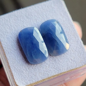 May include: Two faceted, oval-shaped blue gemstones. The gems have a deep, saturated blue color with multiple facets that catch the light. They are displayed on a light gray surface, possibly in a jewelry setting.