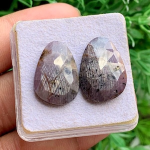 May include: Two pear-shaped, faceted gemstones with a translucent, dusty purple hue. The stones feature visible dark, needle-like inclusions and internal fractures, creating a complex, textured appearance within the polished, reflective surfaces.