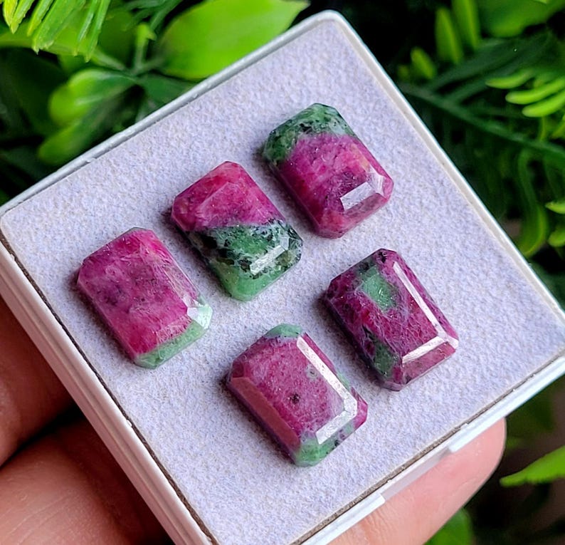 May include: Four rectangular faceted gemstones featuring a mix of vibrant magenta-pink and deep forest green mineral inclusions. The stones are arranged in a display box with a light grey felt lining, showcasing their polished surfaces and edges.