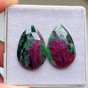 May include: Two pear-shaped faceted gemstones featuring a vibrant mix of deep magenta-red and forest green mineral inclusions. The stones are polished with a reflective surface and are displayed side-by-side on a light gray textured foam insert.