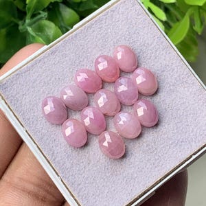 Puede incluir: Un conjunto de doce piedras preciosas rosas de forma ovalada y talladas en facetas, dispuestas en grupo sobre una almohadilla de presentación gris claro texturizada. Cada piedra tiene una superficie pulida con facetas geométricas que reflejan la luz.