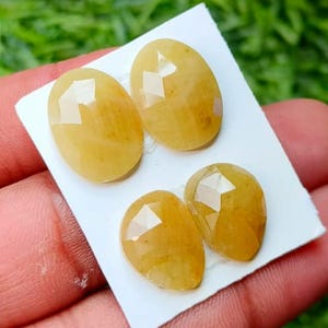 May include: Four faceted yellow gemstones arranged on a white card. Two larger oval-shaped stones are positioned at the top, while two smaller pear-shaped stones are placed below them. Each gemstone features a polished surface with geometric facets.