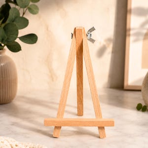 May include: A light-coloured wooden tripod easel featuring three vertical legs connected at the top by a silver-toned metal wing nut and bolt mechanism. The structure includes a horizontal support bar across the front for holding items.
