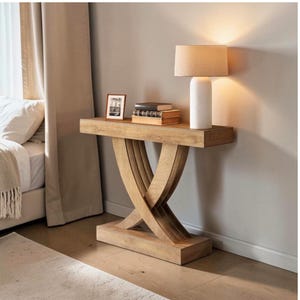 May include: A wooden console table with an X-shaped base. A white lamp with a beige lampshade sits on the table, along with books and a framed picture. The table is in front of a neutral-coloured wall. The table is made of light-coloured wood.