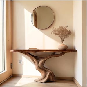 May include: A console table crafted from a single piece of natural wood with a live edge top and a sculpted, tree-trunk-like base. A round mirror hangs on the wall above, and a light-coloured ceramic vase containing dried branches sits on the table.