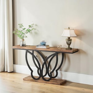 May include: A wooden console table featuring a butterfly-shaped black metal base. The tabletop is a warm brown colour, and the table is decorated with a lamp, books, and decorative objects. The table is placed against a neutral wall.