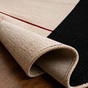 Folded corner of a modern geometric wool rug displaying its flexibility, thickness, edge binding, and handwoven construction in ivory, black, and red tones.