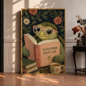 May include: A framed wall art print featuring a green turtle wearing pink glasses, reading an open book with the text SLOW DOWN ENJOY LIFE printed on the cover. The scene includes a stack of books, a floral mug, and a dark background with flowers.