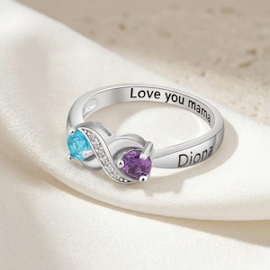Op de afbeelding: Een zilveren ring met een oneindigheidssymboolontwerp, met een blauwe en een paarse edelsteen. De ring is gegraveerd met "Love you mama" en de naam "Diona". De ring ligt op een witte stof.