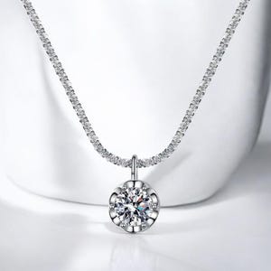 Puede incluir: Un collar de tono plateado con un colgante de piedra redonda, clara y facetada, engastado en un marco de metal festoneado. La cadena está compuesta por pequeños eslabones interconectados que crean un aspecto texturizado y brillante.