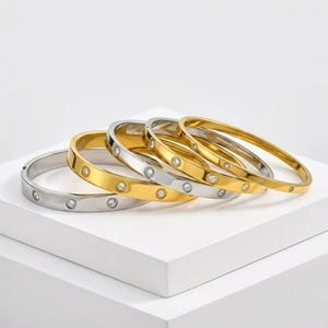 Könnte beinhalten: Fünf gold- und silberfarbene Armreifen mit klaren, runden Edelsteinen. Die Armreifen sind auf einer weißen Oberfläche angeordnet, mit einer Mischung aus Gold- und Silbertönen. Die Edelsteine sind gleichmäßig verteilt.