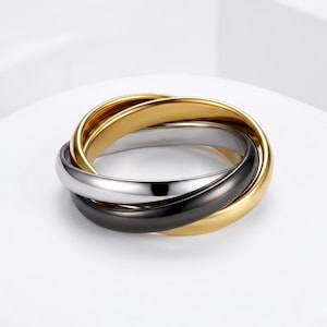 Op de afbeelding: Een armband met drie ringen met in elkaar grijpende banden in goud, zilver en zwart. De banden zijn glad en glanzend, waardoor een moderne en elegante uitstraling ontstaat. De armband is waarschijnlijk gemaakt van roestvrij staal of een vergelijkbaar metaal.