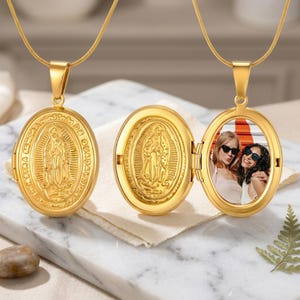Op de afbeelding: Goudkleurige ovale medaillonketting met een gedetailleerde religieuze gravure op de voorkant. Eén medaillon is open en toont een foto van twee personen. De ketting ligt op een marmeren oppervlak.