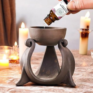 Peut inclure: Un diffuseur d'huiles essentielles marron avec un dessus en forme de bol et des supports incurvés décoratifs. Une main verse de l'huile d'une petite bouteille dans le bol. Des bougies allumées sont en arrière-plan, créant une ambiance chaleureuse.